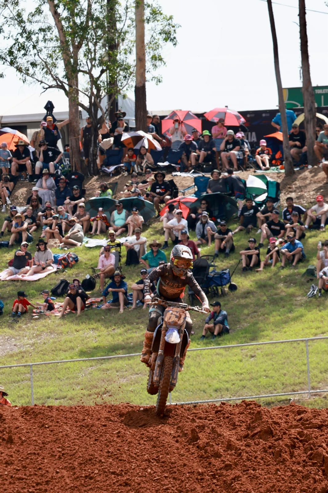 Motocross rider races over the red dirt track with a large crowd of spectators watching from the grassy hillside at MXGP Australia.