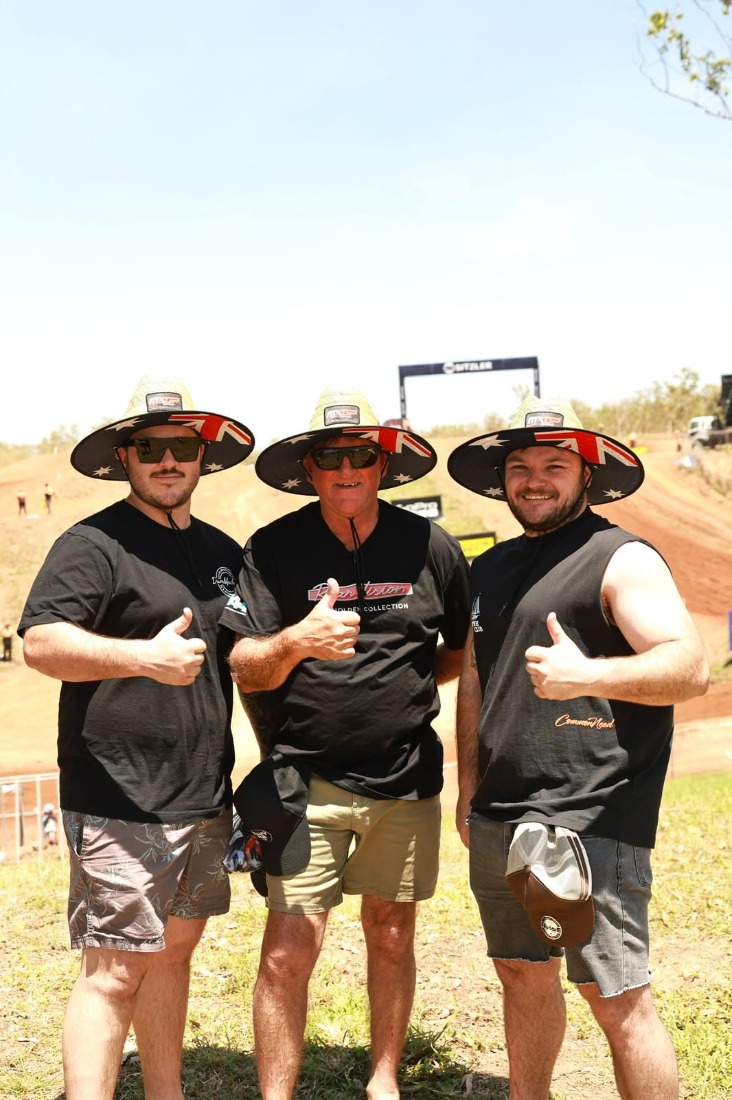 Three MXGP of Australia spectators wearing wide-brimmed Australian flag hats and giving thumbs up beside the motocross track.