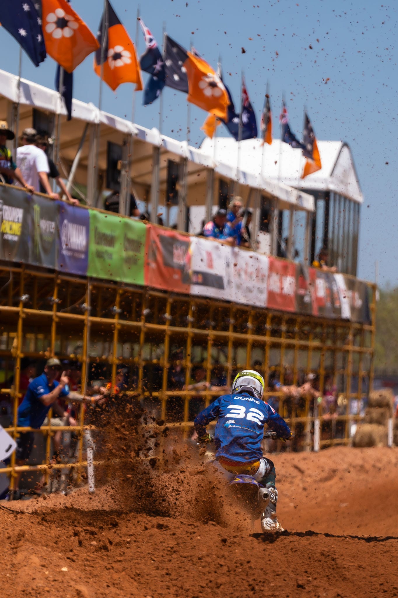Motocross rider number 32 powers through red dirt beside the main spectator structure at MXGP of Australia.
