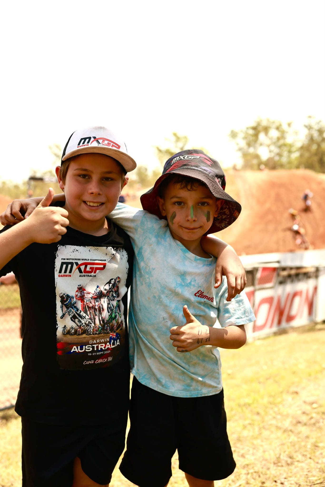 Two young MXGP of Australia spectators wearing event hats pose together and give thumbs up beside the track.