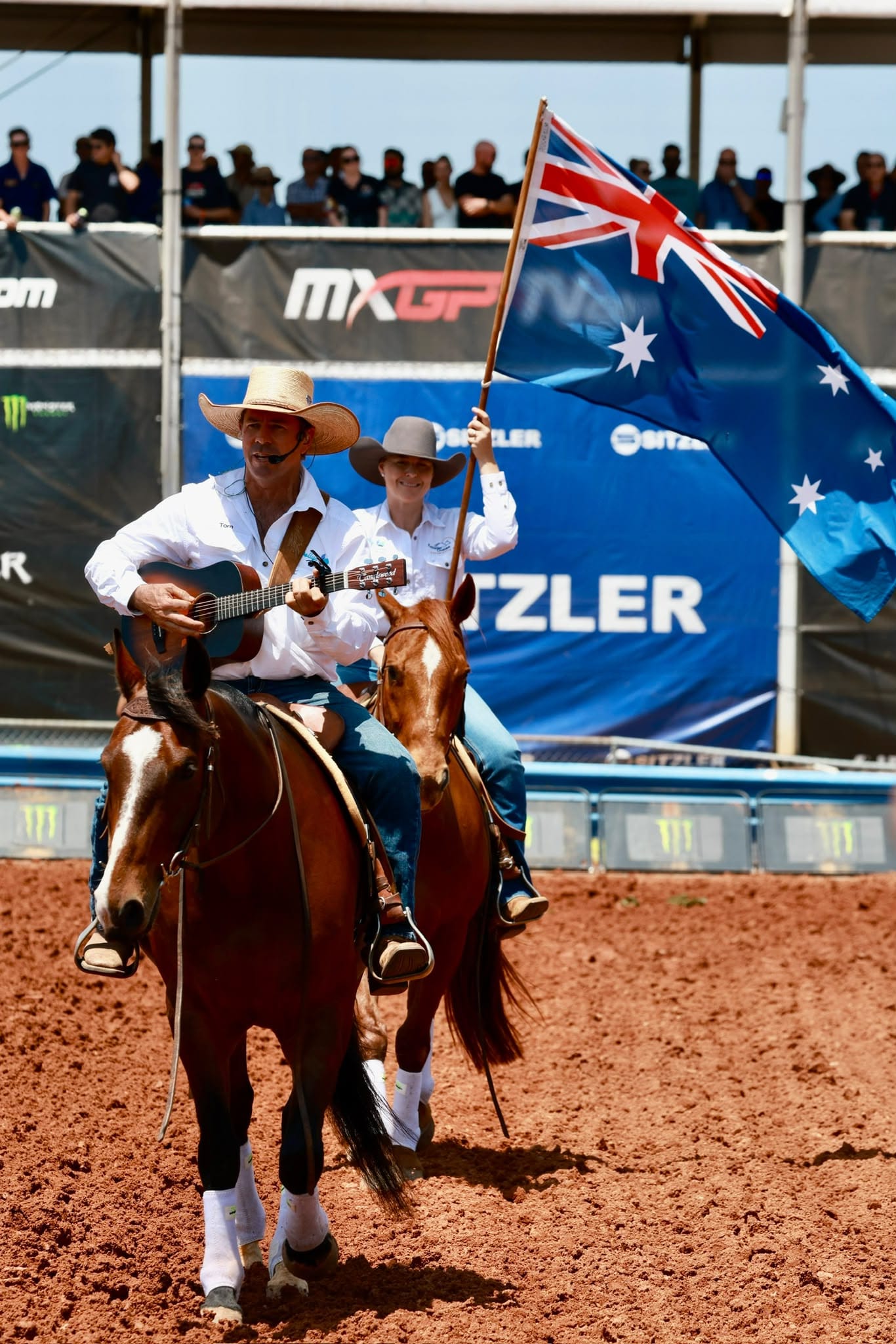 Two riders on horseback enter the track, with one playing guitar and the other carrying a large Australian flag at MXGP Australia in Darwin.