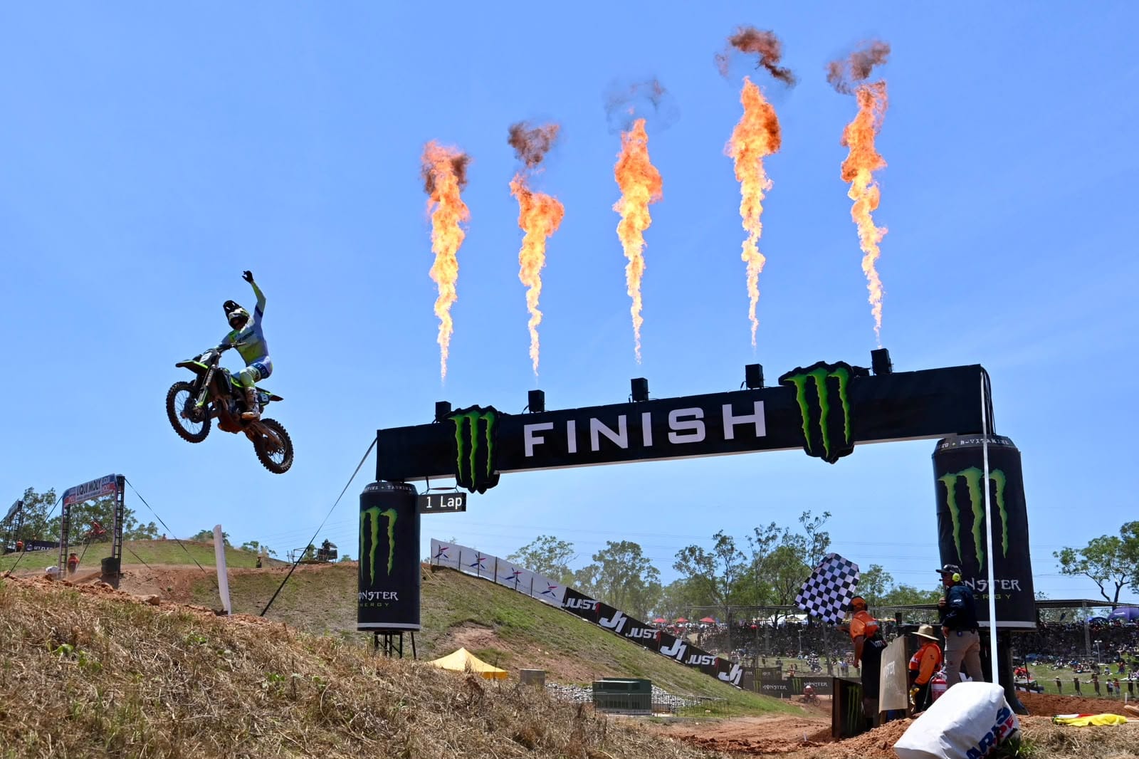 Motocross rider jumps beside the finish line as flames shoot into the air and a marshal waves the chequered flag at MXGP Australia.