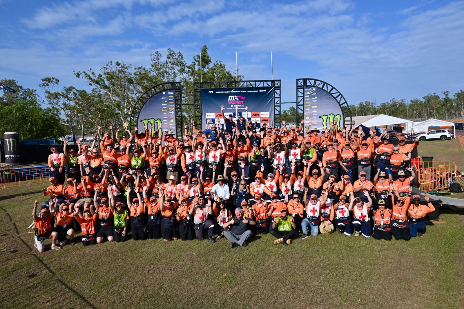 Large group of MXGP Australia officials, marshals, medical staff and event crew pose together with their hands raised in front of the podium at Hidden Valley Raceway in Darwin.