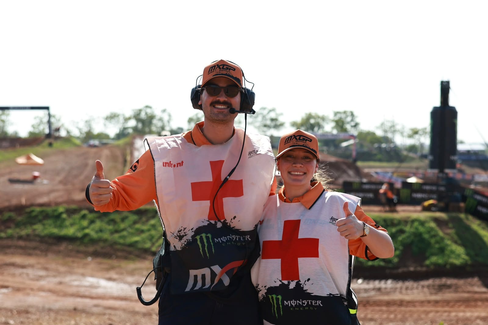 Two MXGP medical staff wearing first aid vests and headsets smile and give thumbs up beside the motocross track in Darwin.
