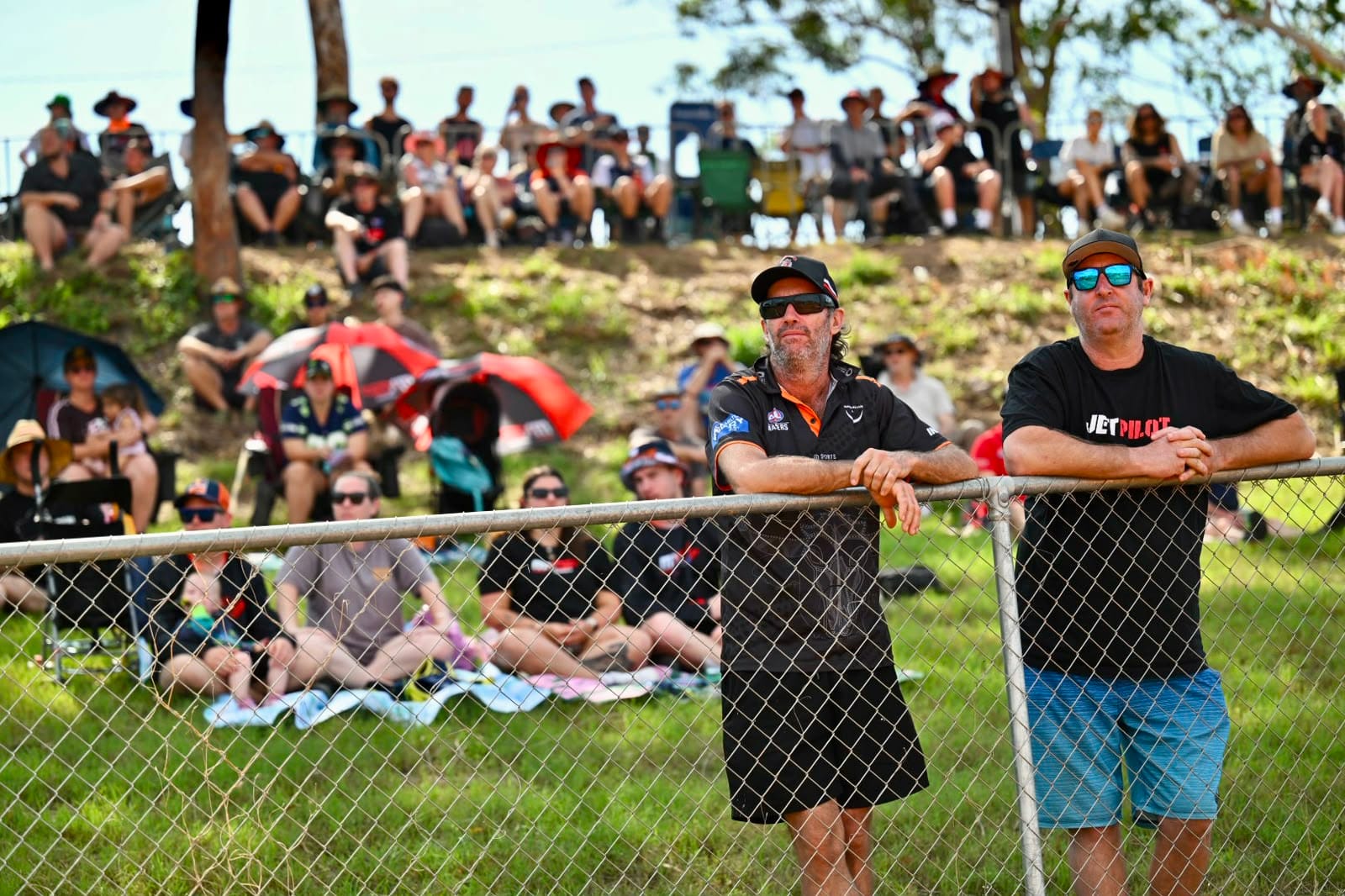 Two spectators lean against a trackside fence while a crowd watches the racing from the grassy hillside behind them at MXGP Australia.