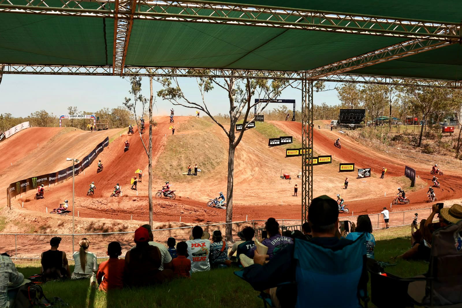 Spectators seated in a shaded viewing area watch motocross riders race around the red dirt track at MXGP of Australia at Hidden Valley.