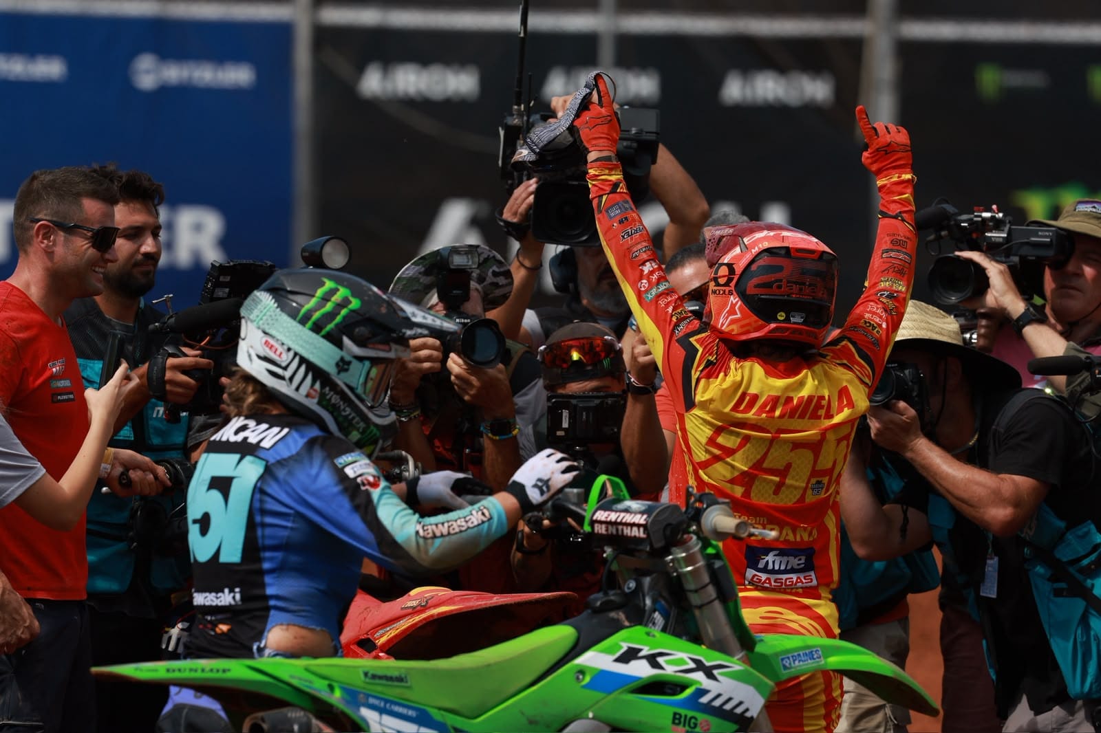 Motocross rider in red and yellow racing gear raises both arms in celebration while surrounded by photographers after the race.