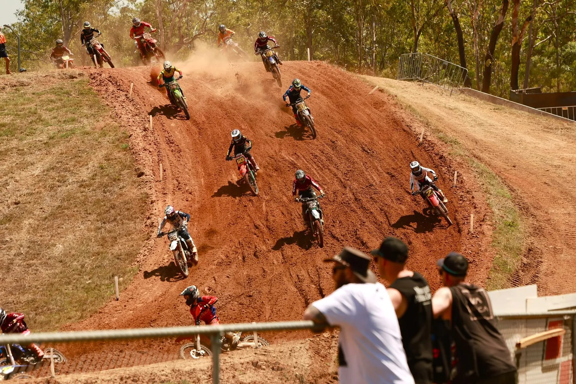 Motocross riders race down a steep red dirt section of the track as spectators watch from nearby.