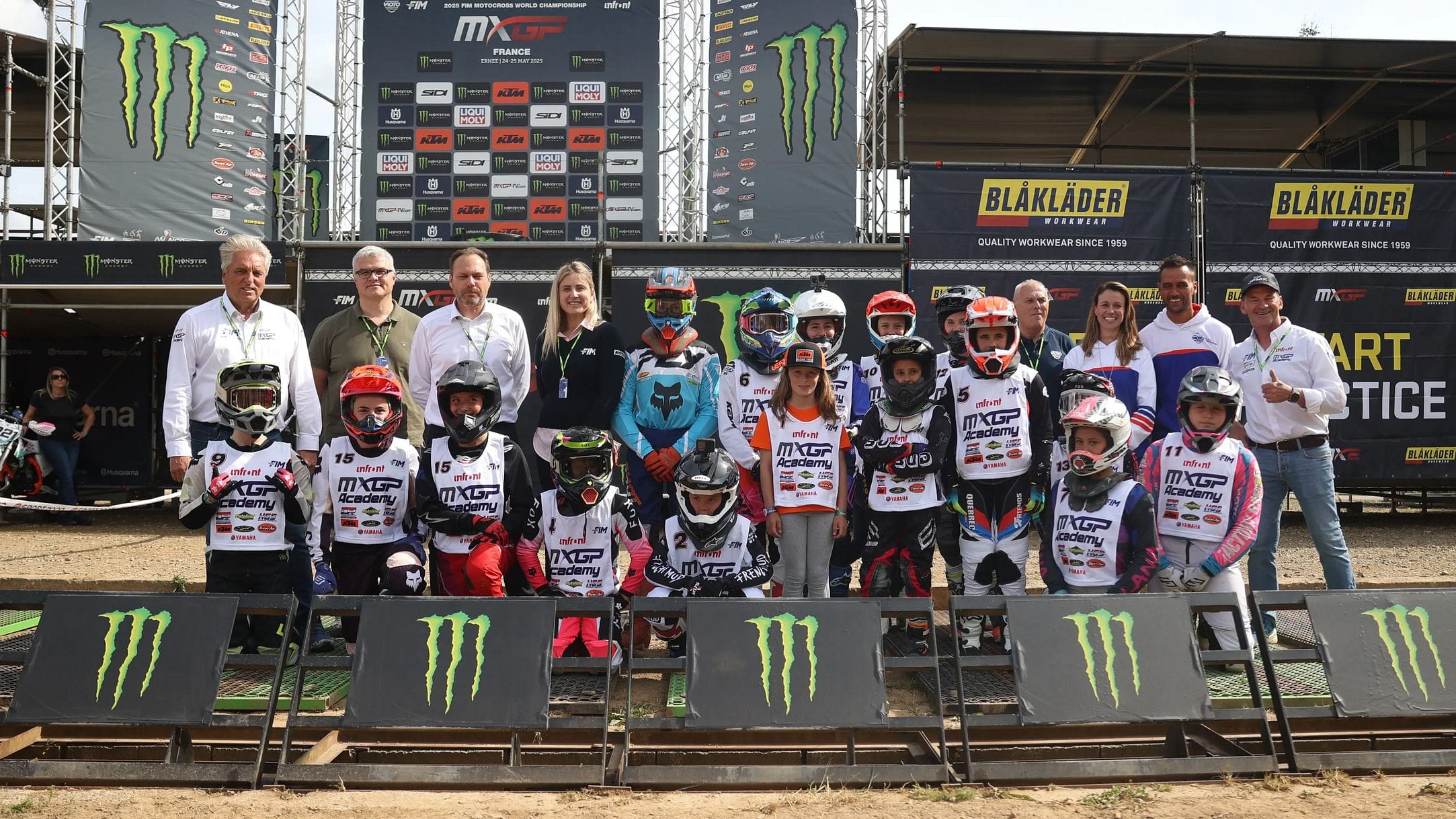 A group of motocross riders in gear and helmets pose with officials in front of a podium at an outdoor event.