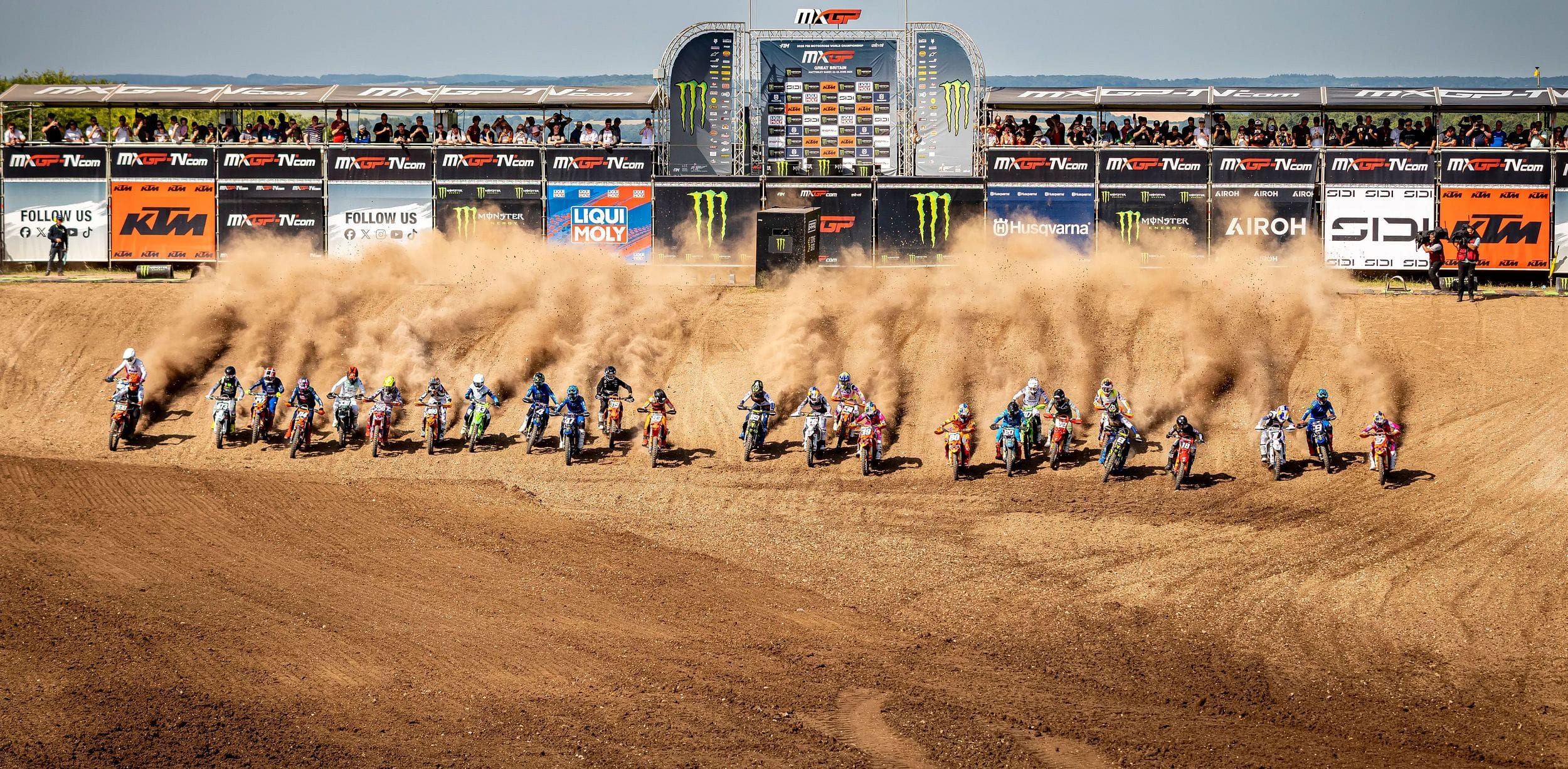 A motocross race starts with multiple riders speeding on a dirt track, kicking up dust. Spectators watch from a platform in the background.