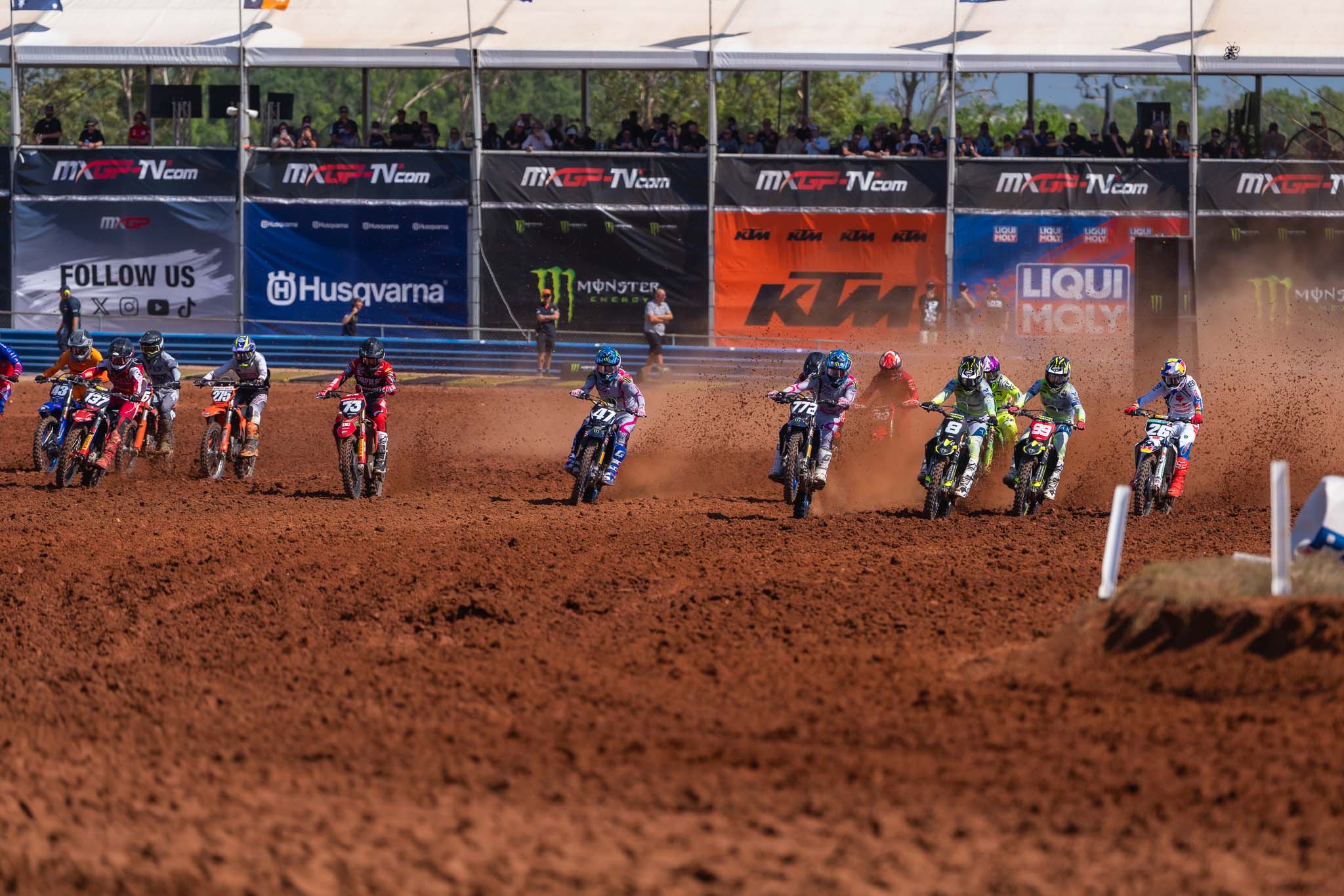 Group of motocross riders race across the red dirt track, kicking up dust as spectators watch from a grandstand at MXGP Australia in Darwin.