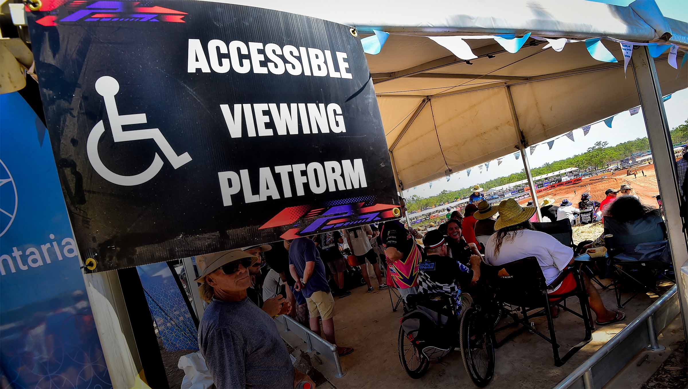 People in wheelchairs under a tent marked "Accessible Viewing Platform" at an outdoor event, with a dirt track visible in the background.